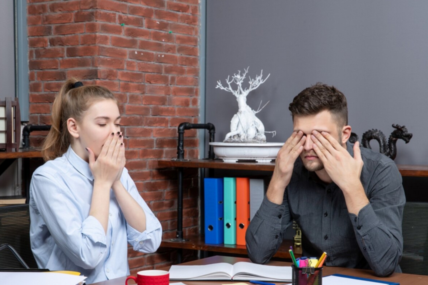 Two coworkers at a desk looking exhausted with hands covering their faces, illustrating signs of burnout and emotional exhaustion