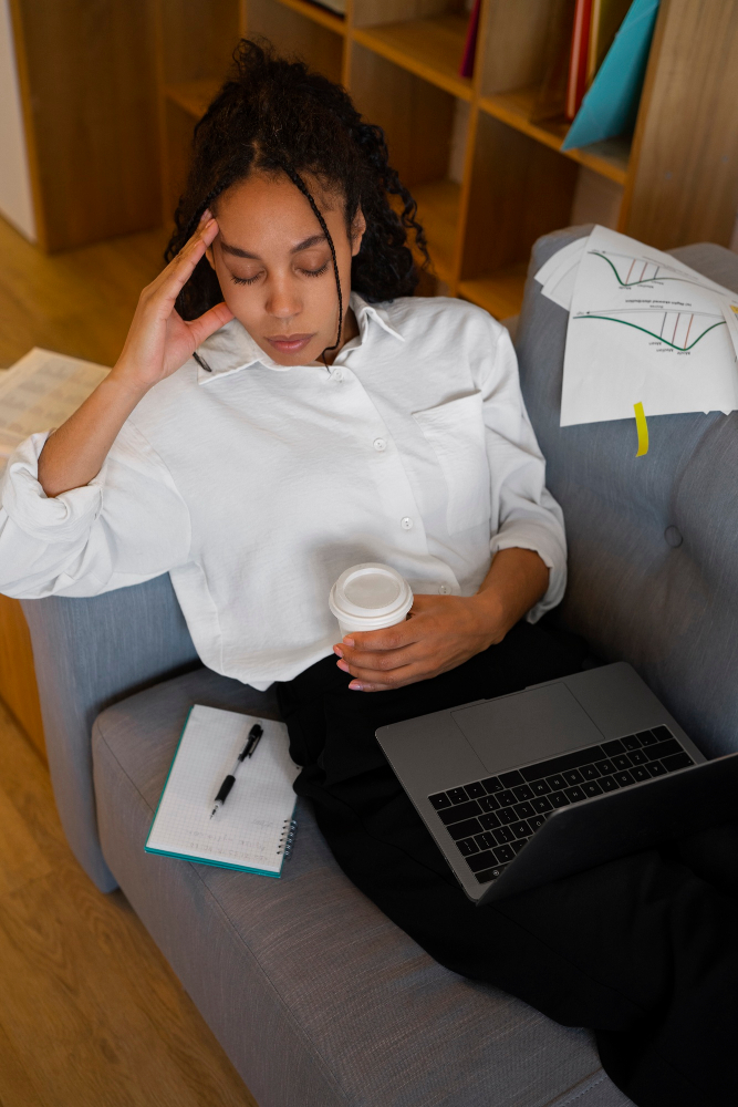 Woman sitting on a couch with a laptop and coffee, hand to her head looking stressed, representing mental health challenges