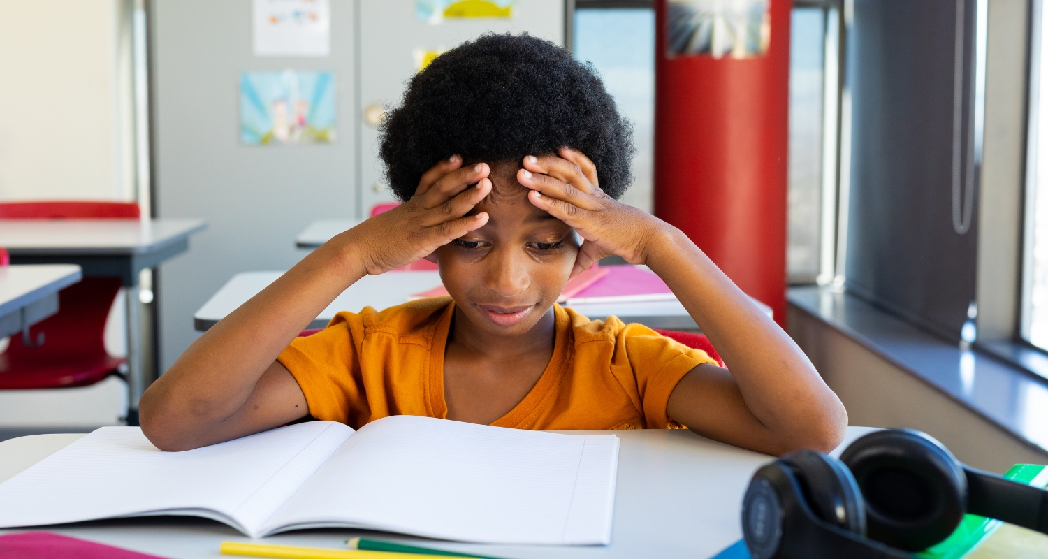 Child at school desk with hands on head looking frustrated, representing ADHD challenges in focus and attention