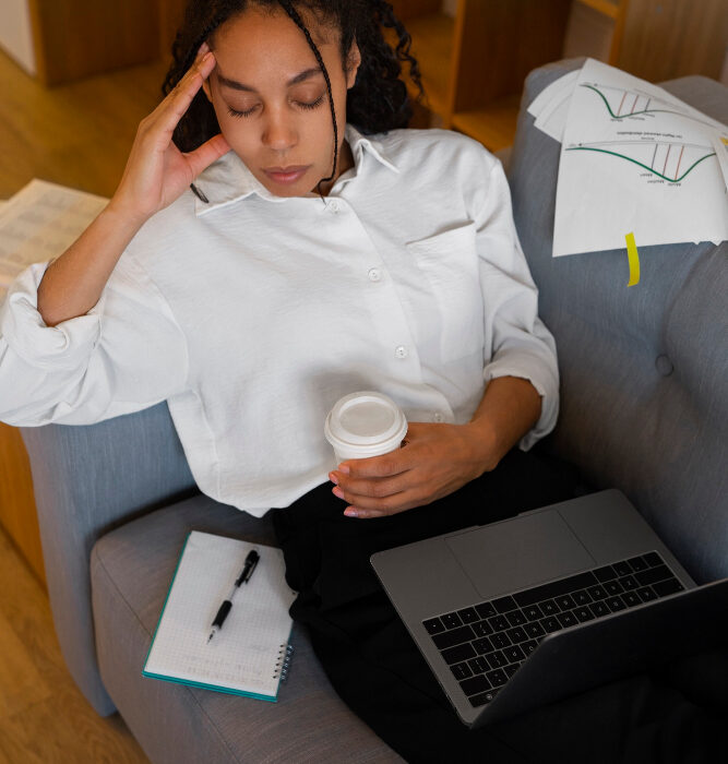 Woman sitting on a couch with a laptop and coffee, hand to her head looking stressed, representing mental health challenges