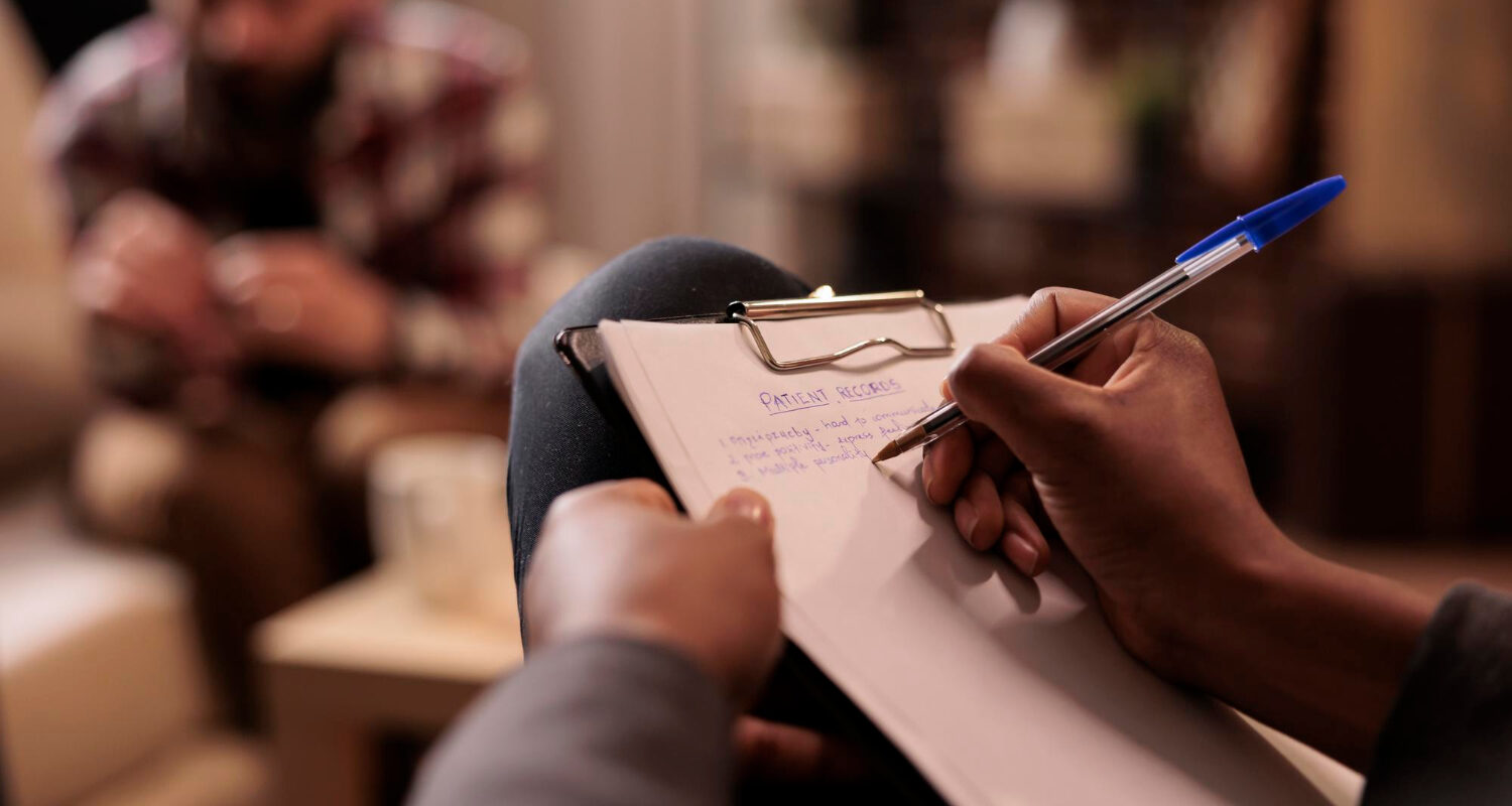 Clinician writing notes on a clipboard during a psychiatric evaluation with a patient in the background