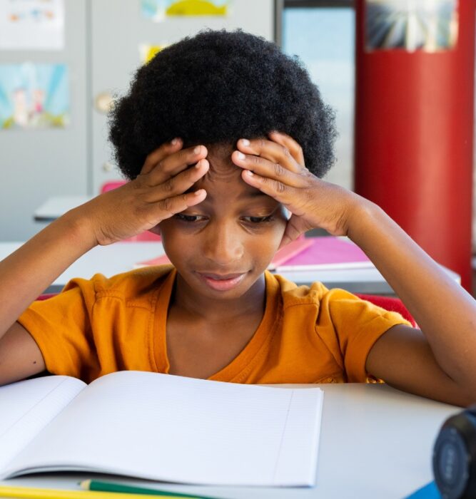Child at school desk with hands on head looking frustrated, representing ADHD challenges in focus and attention